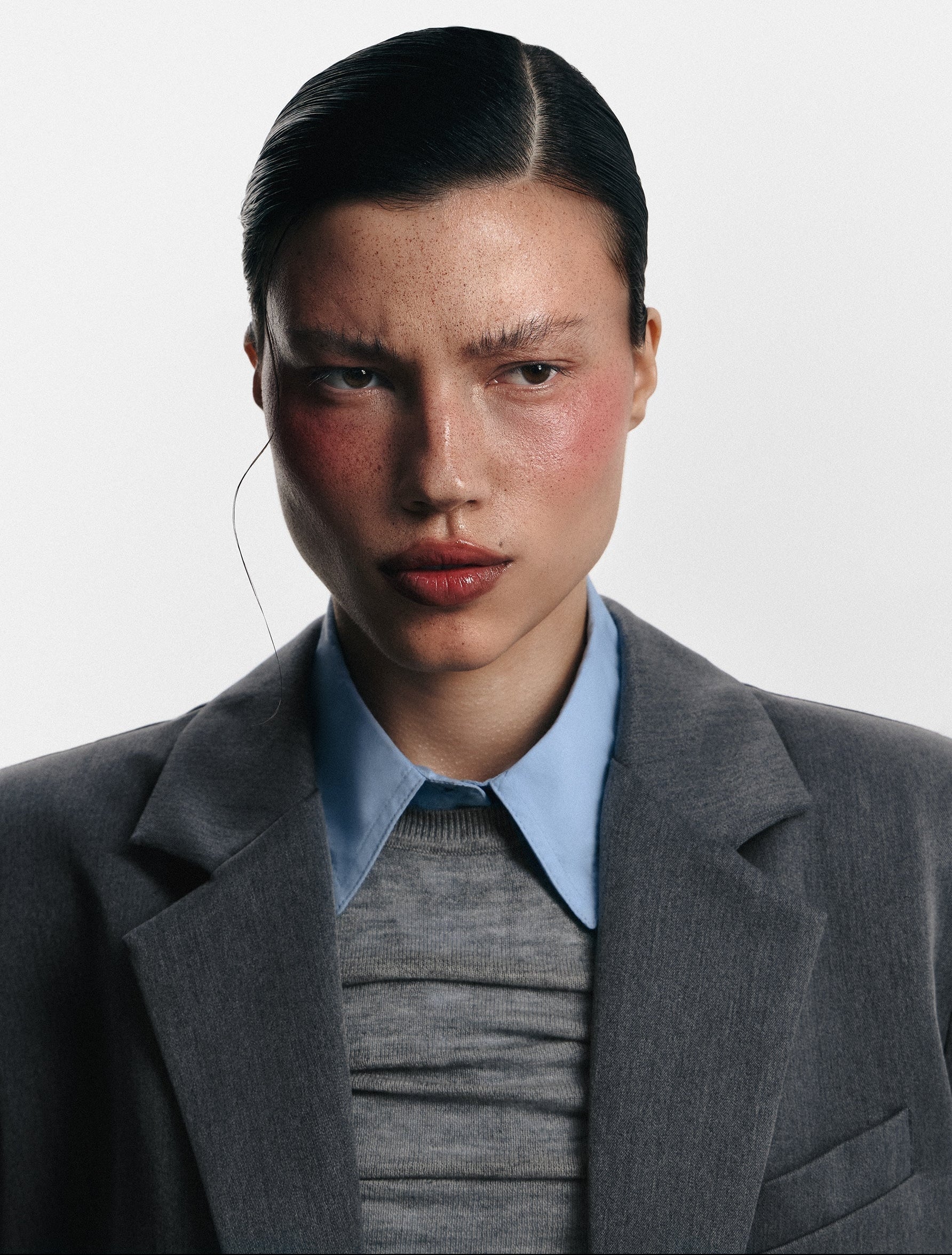 A close-up portrait of a model wearing a structured gray blazer over a light blue collared shirt and a gray ribbed sweater, against a plain light gray background.